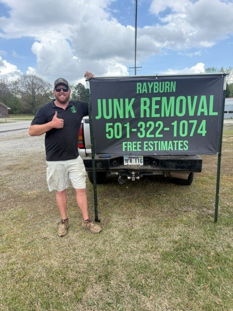 Rayburn Junk Removal team member beside branded signage, smiling with a thumbs up